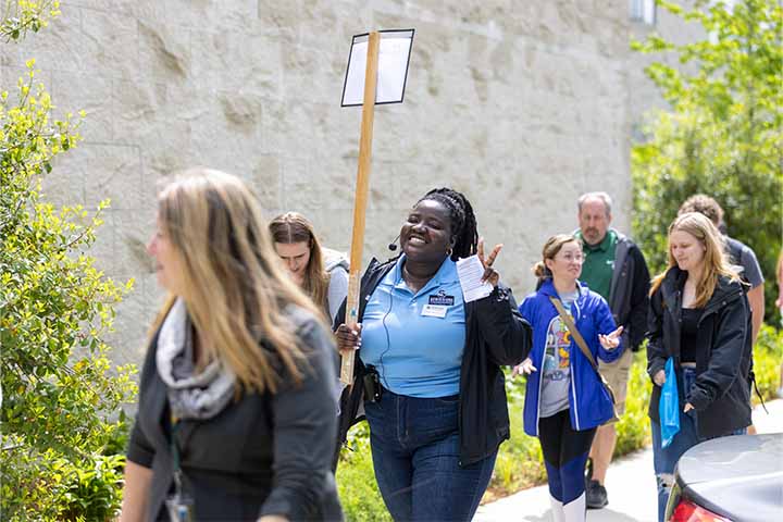 An Admissions Ambassadors holds a tour sign and gives a peace sign with her hands