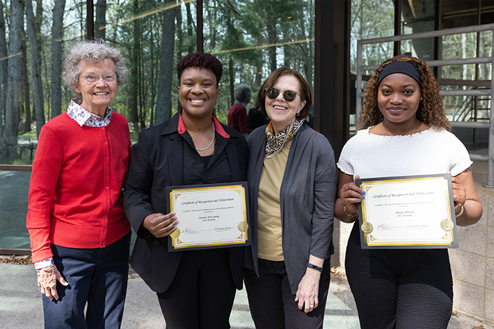 Four women pose outside Stockton's lakeside lodge; two who are students hold their award certificates