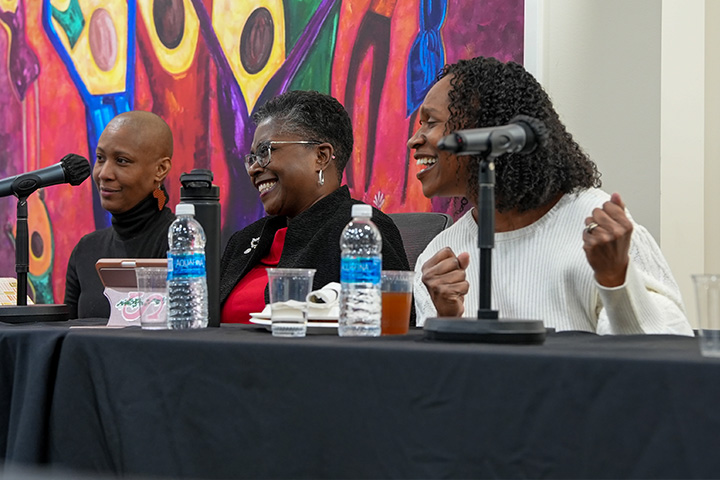 Three panelists sit at a long table with tall microphones with colorful artwork on the wall behind them