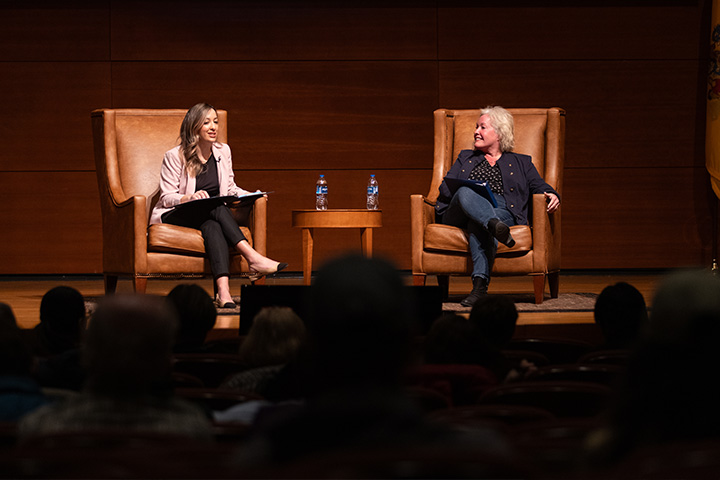 Two women sit in large brown leather chairs on stage in the Campus Center Theatre