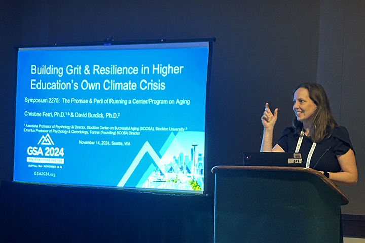 Christine Ferri stands behind a podium next to a screen presenting a PowerPoint presentation