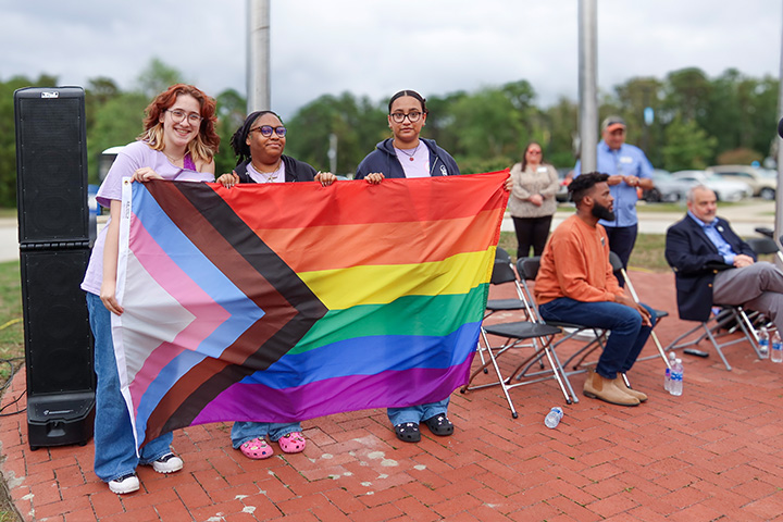 Three students hold the pride flag, which combines the rainbow horizontal stripes with black, brown, blue, pink and white chevron stripes 