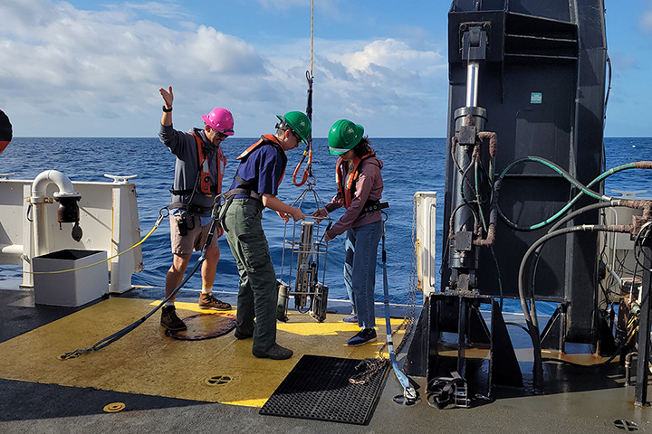 Students in hard hats and harnesses examine equipment on a boat with the wide blue ocean behind them