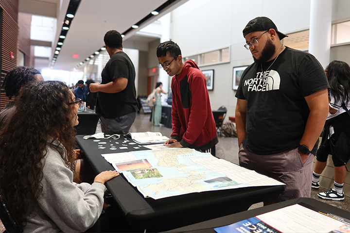 A student seated behind a table chats with two students standing on the other side at the Study Abroad Fair