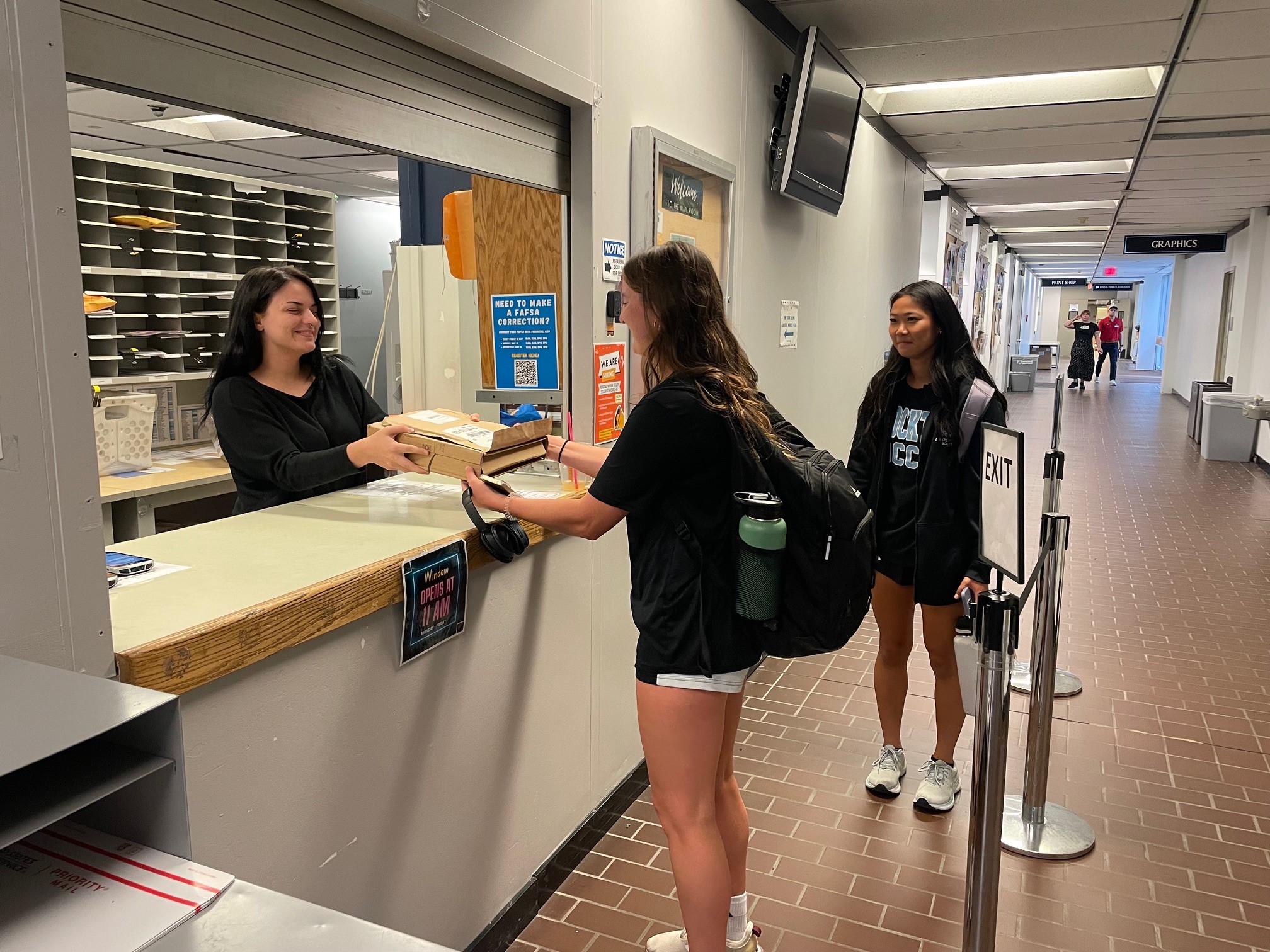 Two students in line at the mailroom window. One student is working and handing over the package to the recipient student. Two students in line at the mailroom window. One student is working and handing over the package to the recipient student.