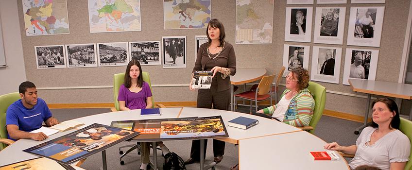 Student sitting around a table in the Holocaust Resource Center, with a professor showing books and photographs.