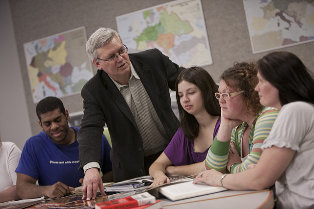 Students sitting around a table while a faculty member shows them documents. Maps are hung on the wall in the background.